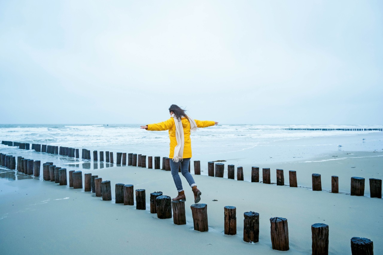 Vrouw met gele jas op paaltjes bij de zee
