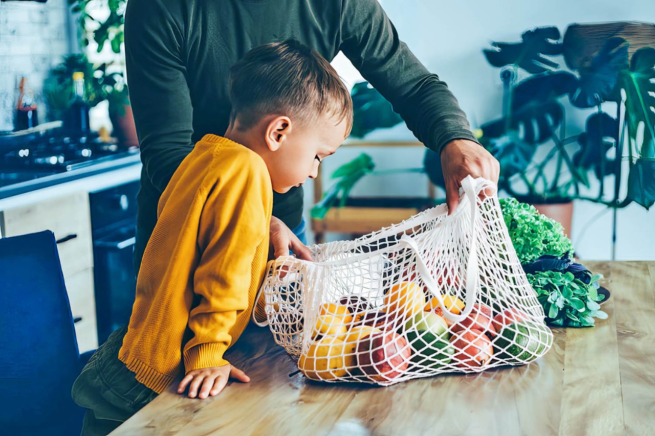 Jongetje met gele trui kijkt in een boodschappennet met fruit op tafel
