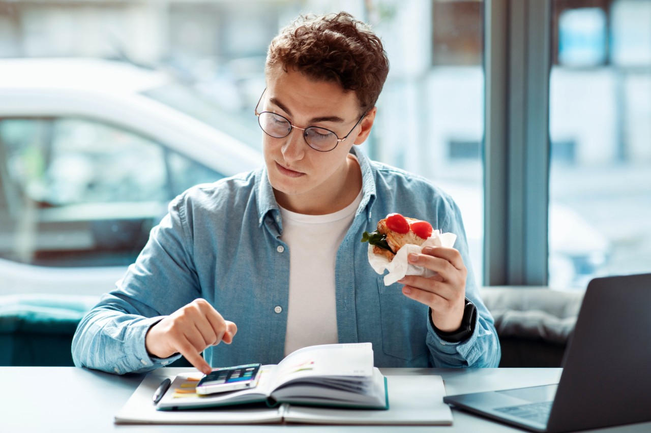 Curly-haired caucasian young man student with glasses multitasking by eating a healthy wrap and checking his calculator over an open planner near his laptop in a cafe. Work, study with food
